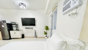 A hotel room with a bed, a TV mounted on the wall, a refrigerator, a coffee machine, a small cabinet, and a potted plant.