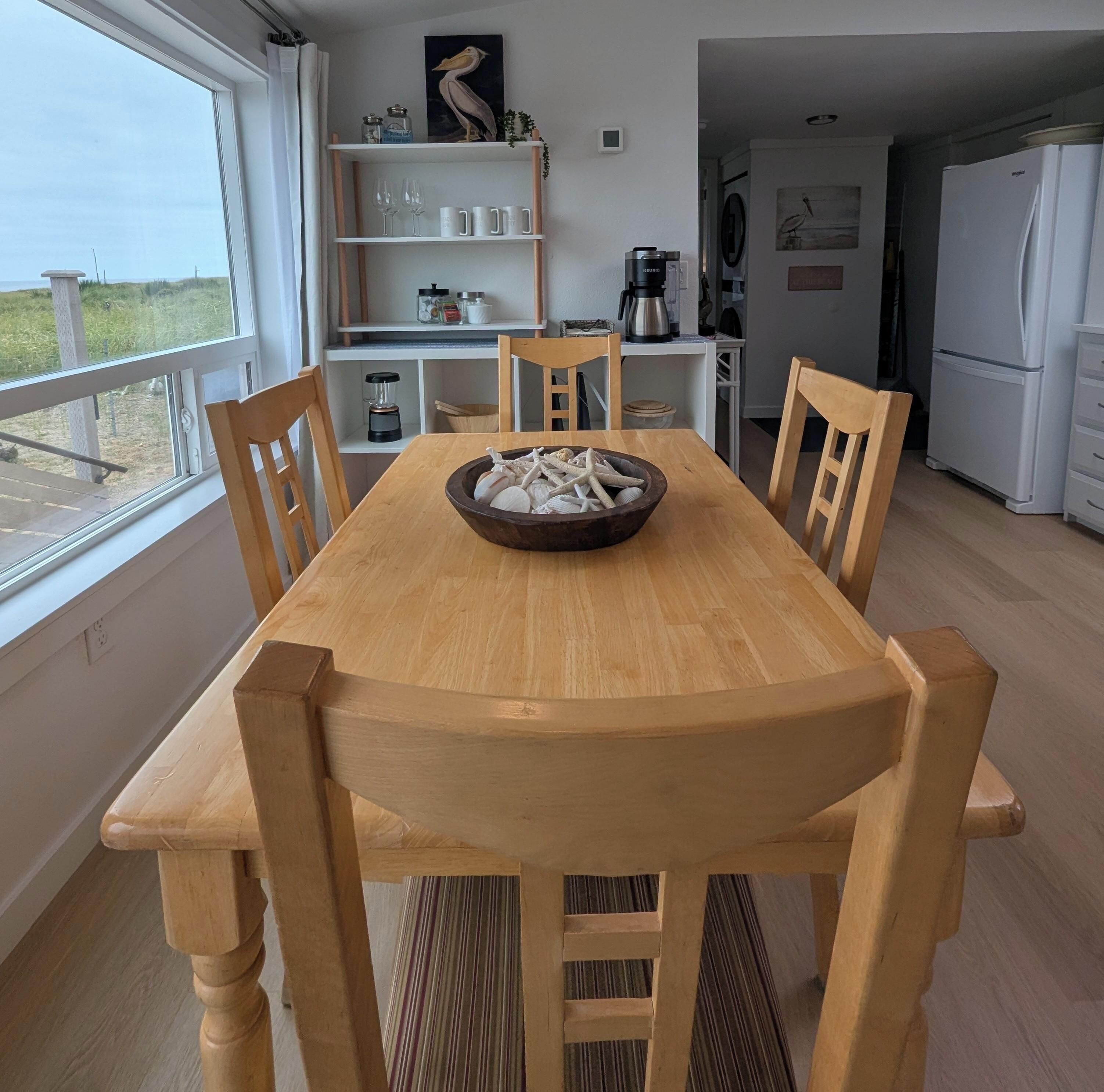 Comedor con mesa y sillas de madera, un cuenco de estrellas de mar y vistas al exterior a través de un ventanal.