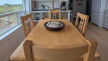 Comedor con mesa y sillas de madera, un cuenco de estrellas de mar y vistas al exterior a través de un ventanal.