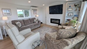 A living room with a grey sofa, a white armchair, a patterned armchair, a fireplace, and a TV mounted on the wall.