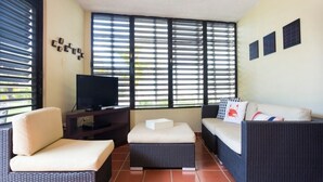 A living room with a television, two sofas, a coffee table, and large windows with blinds.
