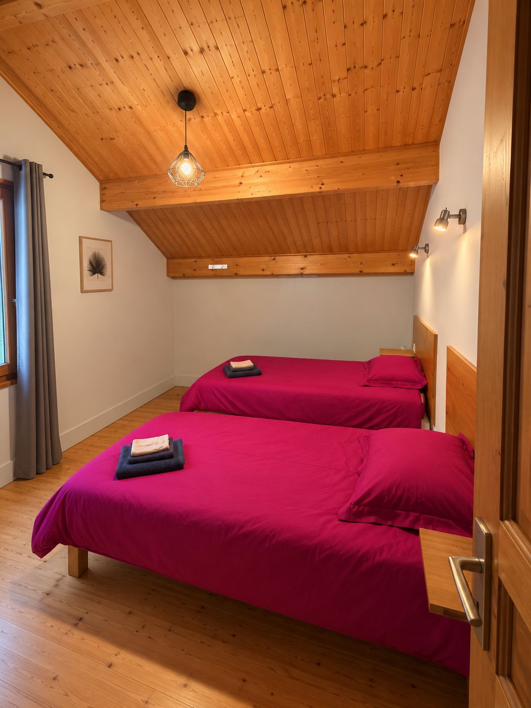 Una habitación con dos camas, suelo de madera, techo inclinado con vigas de madera, una lámpara colgante y un cuadro en la pared.