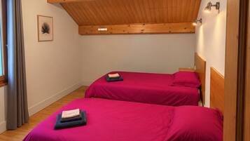 Una habitación con dos camas, suelo de madera, techo inclinado con vigas de madera, una lámpara colgante y un cuadro en la pared.