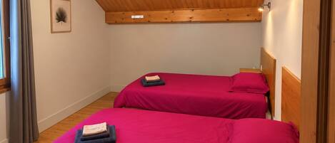 A room with two beds, wooden flooring, a sloped ceiling with wooden beams, a pendant light, and a framed picture on the wall.