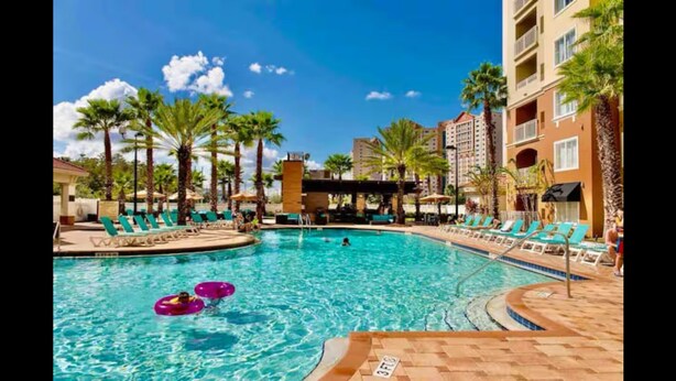A swimming pool with lounge chairs and palm trees, surrounded by hotel buildings.
