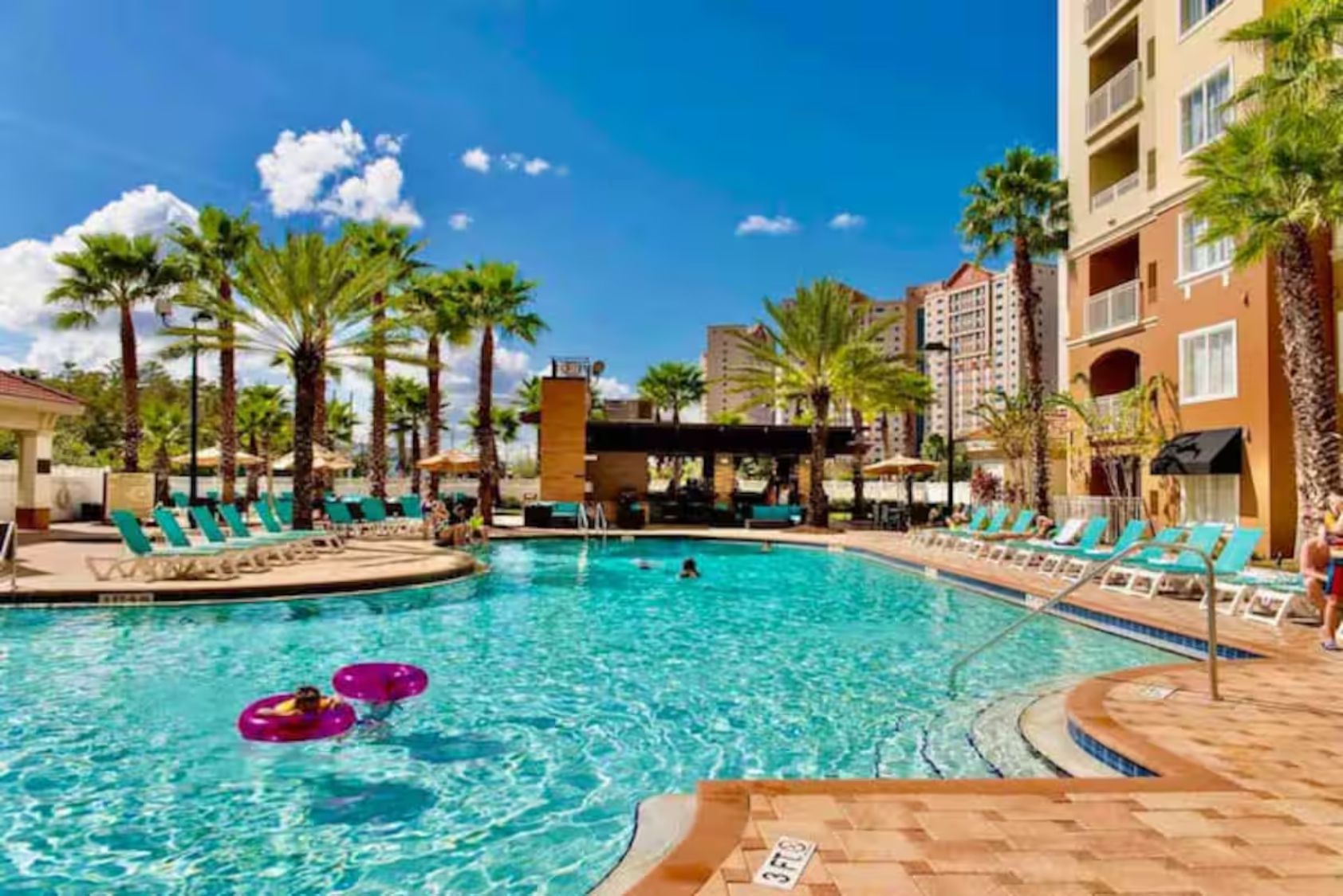 A swimming pool with lounge chairs and palm trees, surrounded by hotel buildings.