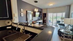 A modern hotel room with a kitchen area, a large window with a view, and a seating area with a sofa and a side table.