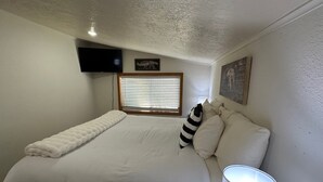 A bedroom with a bed, a television mounted on the wall, a window with blinds, and a painting on the wall.