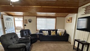 A cozy living room with a dark leather sofa, a matching armchair, and a small coffee table. There is a television mounted on the wall and a door leading outside.