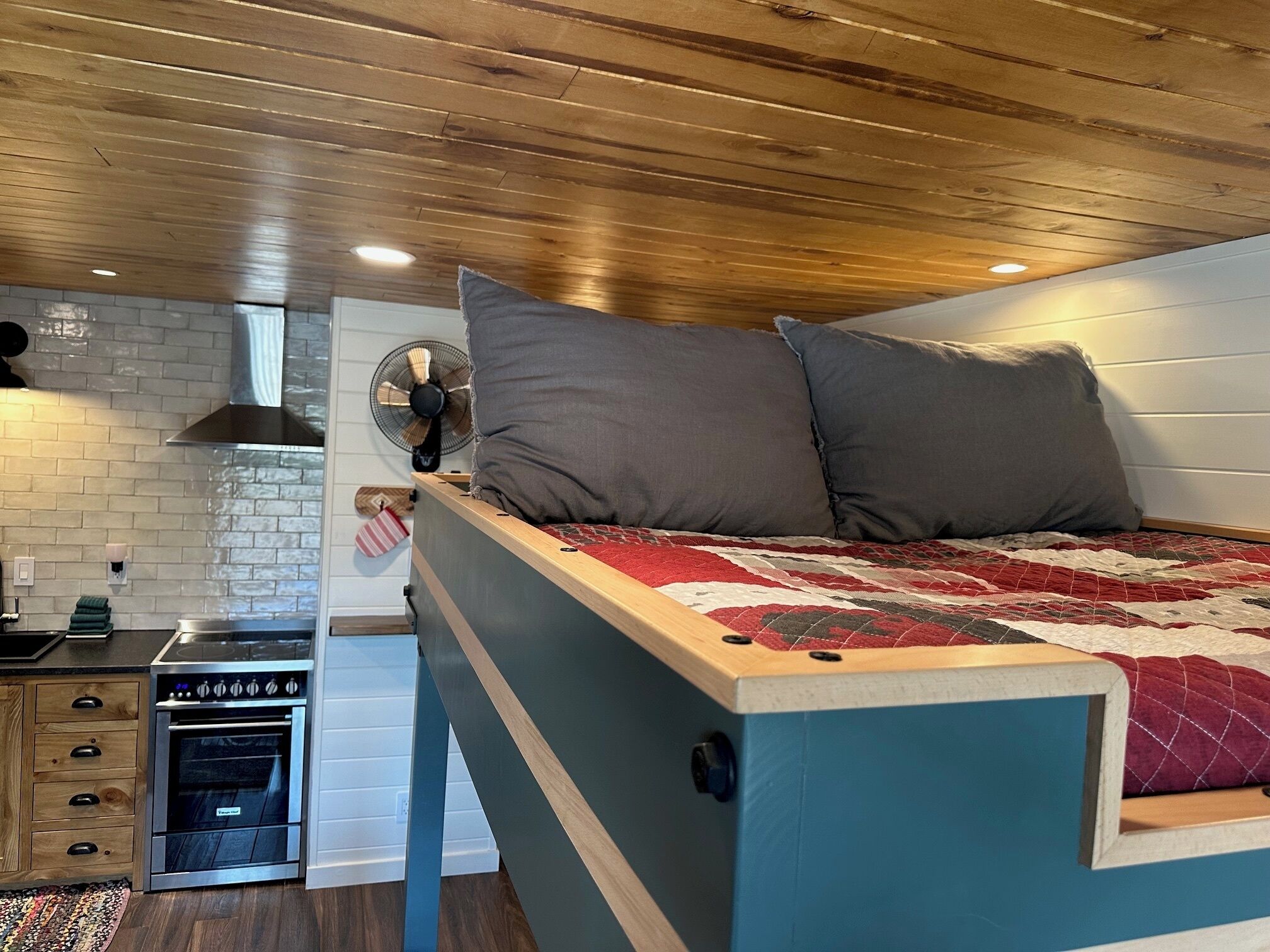 A compact bedroom with a bed, a small kitchenette, and a wooden ceiling.