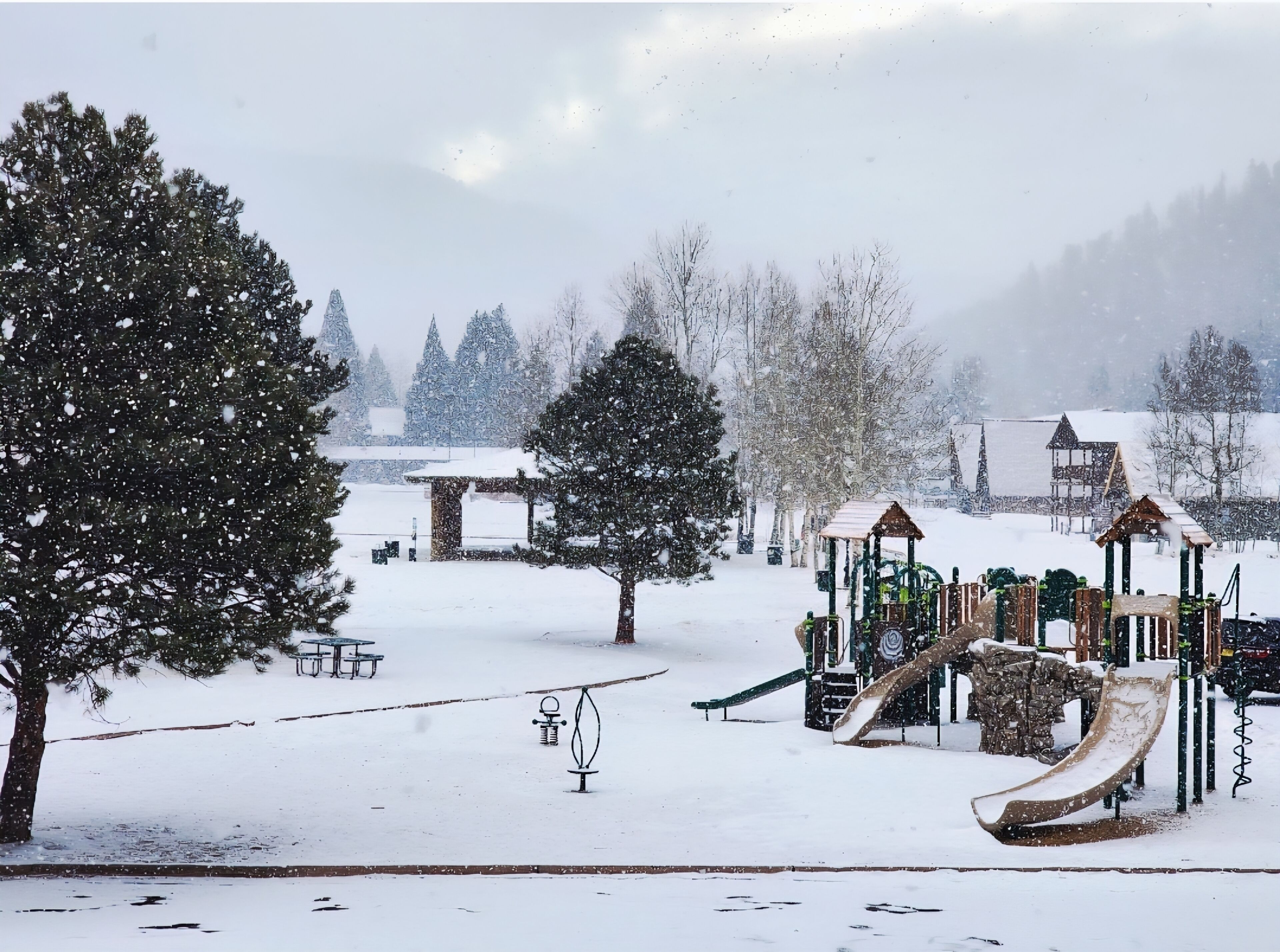 A snow-covered playground with slides and swings, surrounded by trees and a building in the background.