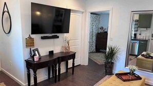 Ein Wohnzimmer mit einem an der Wand befestigten Flachbildfernseher, einem dunklen Holztisch, einem beige gefärbten Sofa und einer Topfpflanze.
