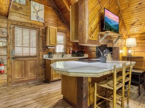 Interior rústico de una cabaña de madera con cocina que incluye una isla grande, armarios de madera, chimenea y televisor montado en la pared.