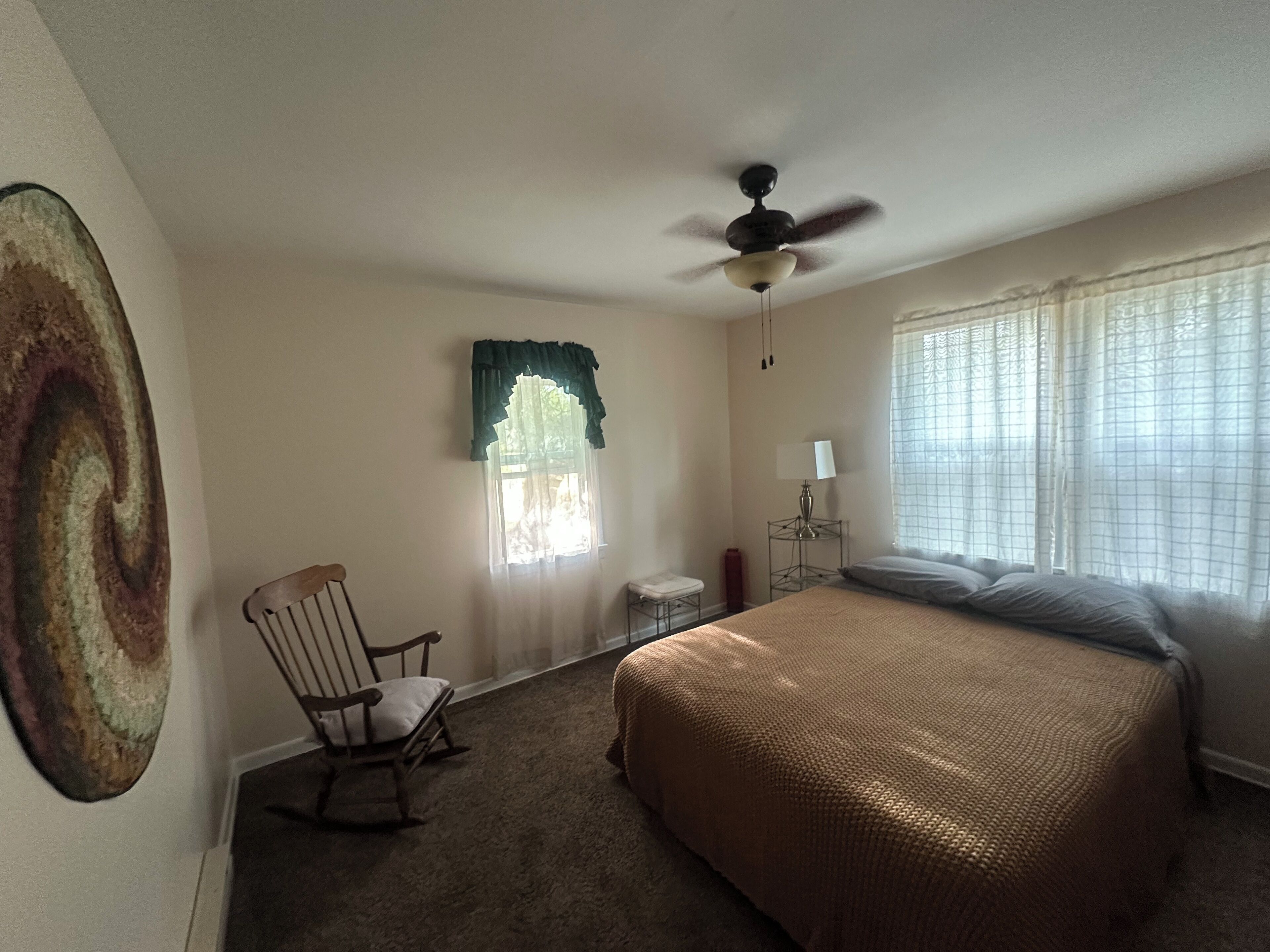 A bedroom with a bed, a chair, a ceiling fan, a window with curtains, and a lamp on a bedside table.