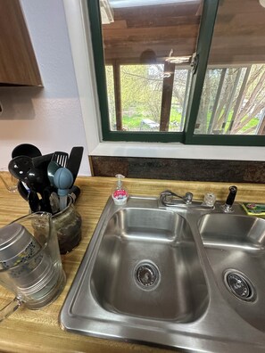 A kitchen with a stainless steel sink, a window with a view of greenery, and various kitchen utensils.