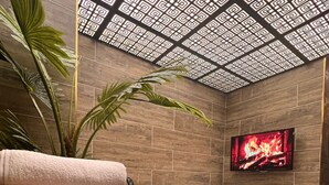 A modern bathroom with a large bathtub, wooden shelves with towels, a potted plant, and a TV displaying a fireplace. - Petra Otel (Eskişehir)