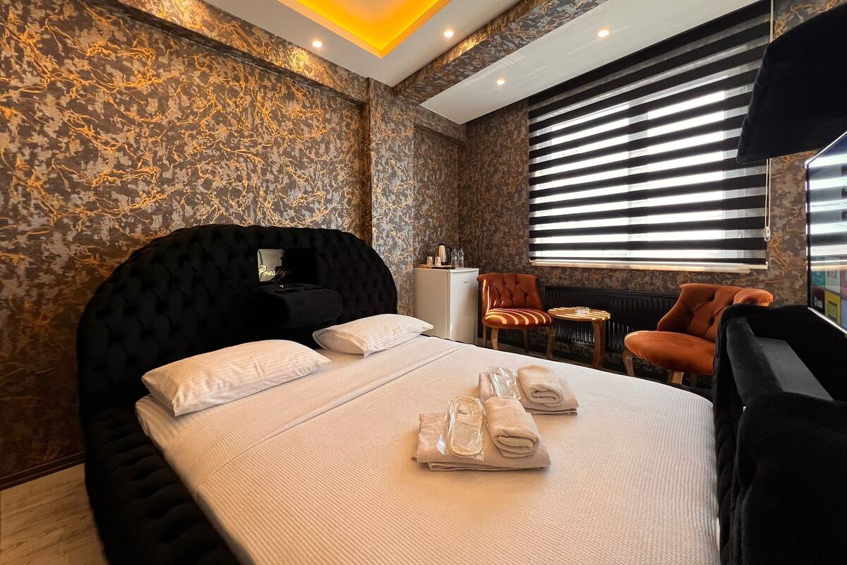 Ein modernes Hotelzimmer mit einem großen Bett, einem Kopfteil, einem kleinen Tisch, zwei Stühlen und einem Fenster mit Jalousien.
