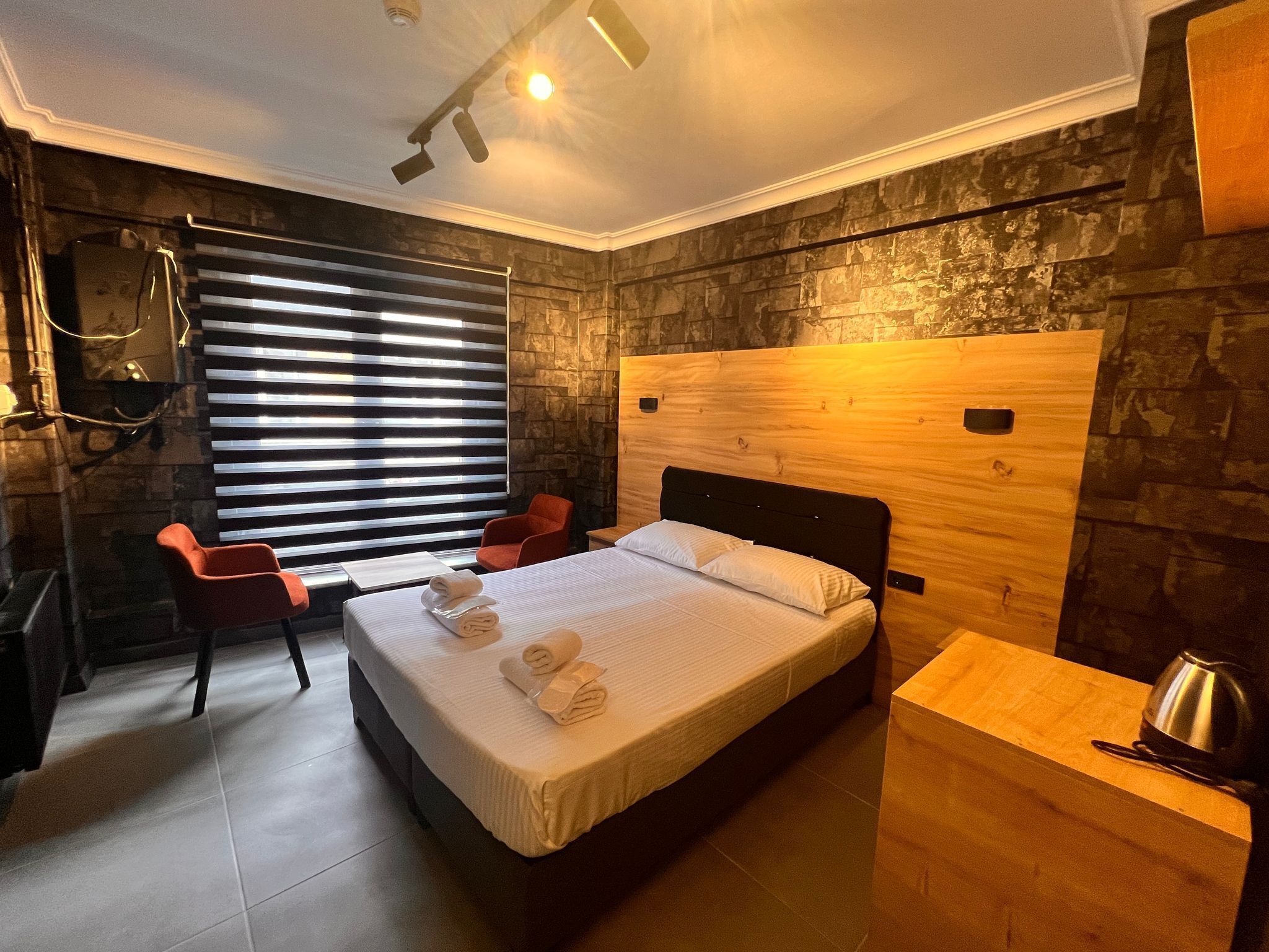 A modern hotel room with a large bed, a wooden headboard, a red chair, a small table with a kettle, and a stone wall.