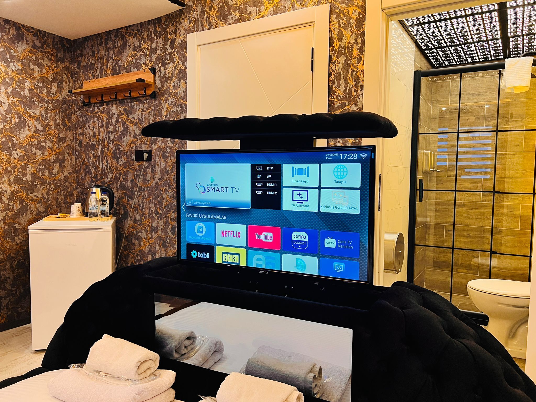 A bathroom with a digital screen displaying various apps, a towel rack, a toilet, and a shower area.