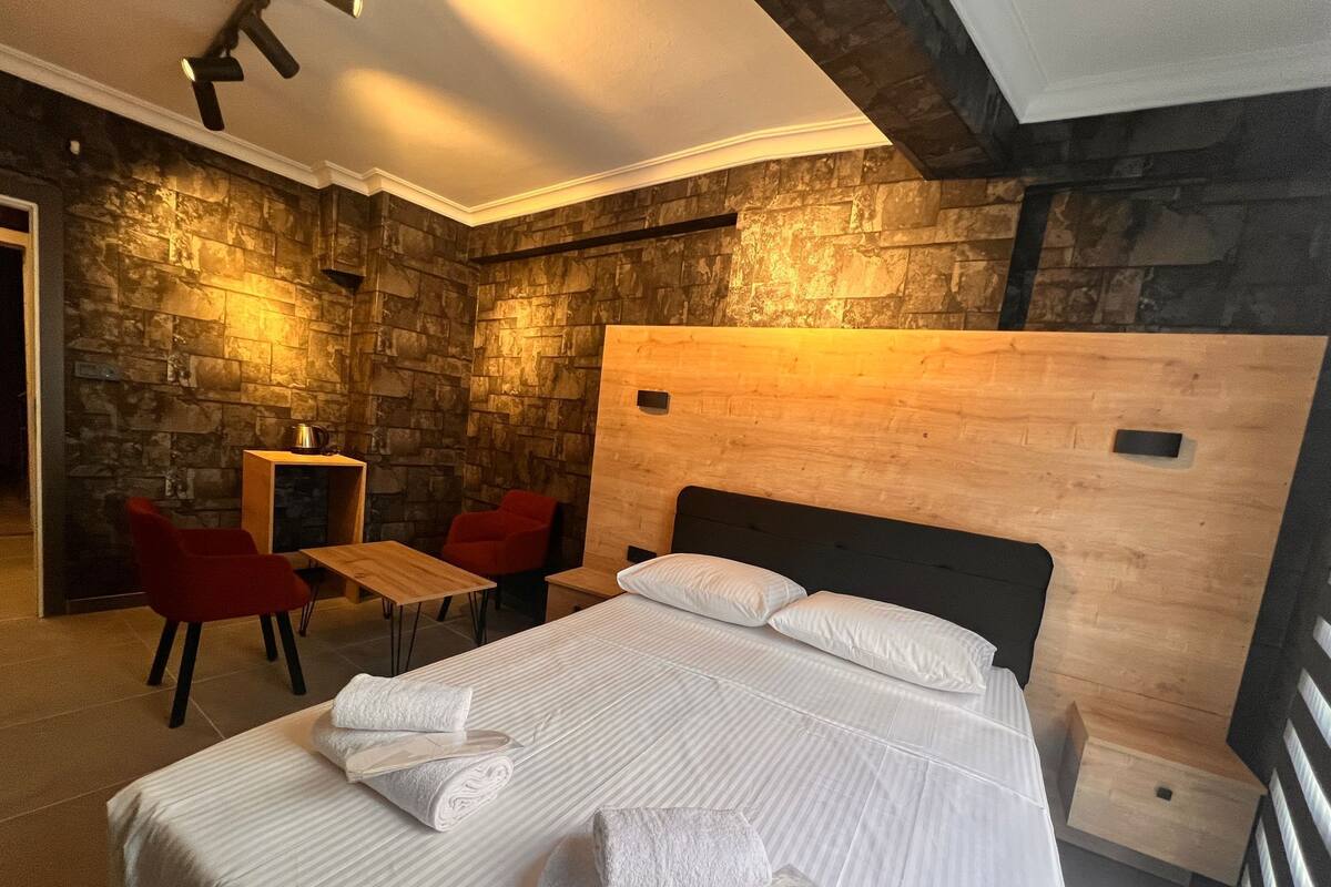 Ein Hotelzimmer mit Stehwänden, einem Bett mit weißer Bettwäsche, einem hölzernen Kopfteil, einem kleinen Tisch und roten Stühlen.