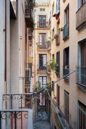Un callejón urbano estrecho flanqueado por edificios, con balcones con plantas y mesas al aire libre.