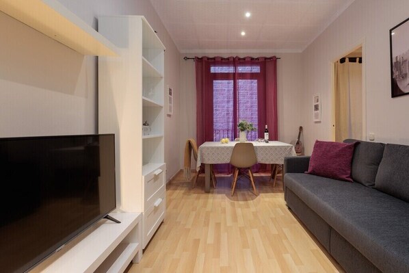 Una sala de estar con televisión, un sofá, una mesa de comedor con sillas y una ventana con cortinas.
