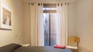 Un dormitorio con cama, mesita de noche, una silla con manta, y vista a un edificio a través de una ventana.