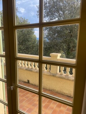 A room with a view of a balcony, a tiled floor, and a window with a view of trees and a clear sky.