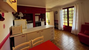 A compact kitchen with a dining area, a red sofa, and a large window with curtains. - House La Baixada (Oms)