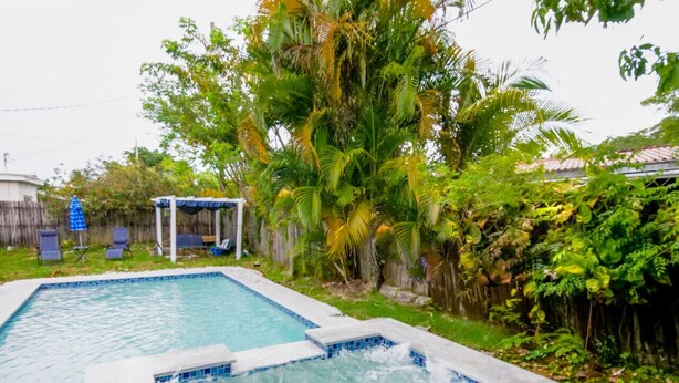 A rectangular swimming pool with a hot tub in the center, surrounded by lush greenery and a wooden fence. - The Blue 4Bedroom Home– Pool & Spa Retreat (North Miami Beach)