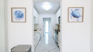 A narrow hallway with a kitchen on the right, framed artwork on the walls, and a trash bin on the left. - The Blue 4Bedroom Home– Pool & Spa Retreat (North Miami Beach)