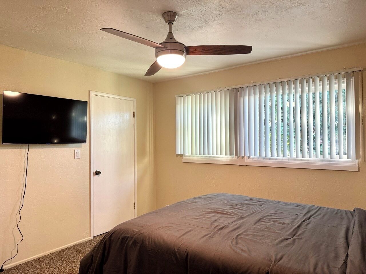 Une chambre dotée d’un ventilateur de plafond, d’un lit, d’un téléviseur fixé au mur et d’une fenêtre avec des stores.