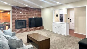 A living room with a brick fireplace, a flat-screen TV, a sofa, and a small wooden coffee table. - SpaciousHome with King Bed, Game Room & Washer/Dryer (Hanford)