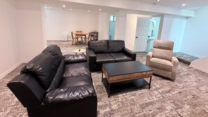 Un salon avec un canapé en cuir noir, un fauteuil beige et une table basse en bois. On aperçoit une table à manger avec des chaises au fond.
