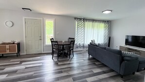 Un salon avec un canapé gris, une table à manger avec des chaises, un téléviseur et une fenêtre avec des rideaux.