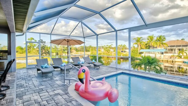 A pool area with lounge chairs, a flamingo float, and a view of palm trees and a lake.