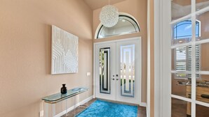A modern entryway with a glass console table, a blue rug, a decorative wall piece, and a large arched window.