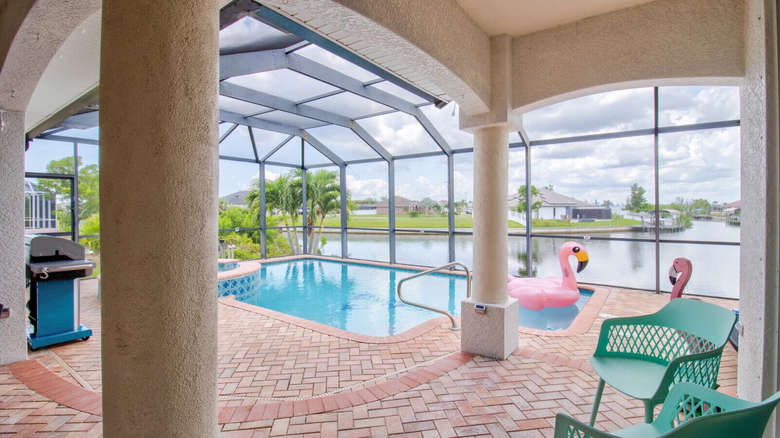 A covered outdoor swimming pool with a view of a lake, a flamingo float, and a grill area.