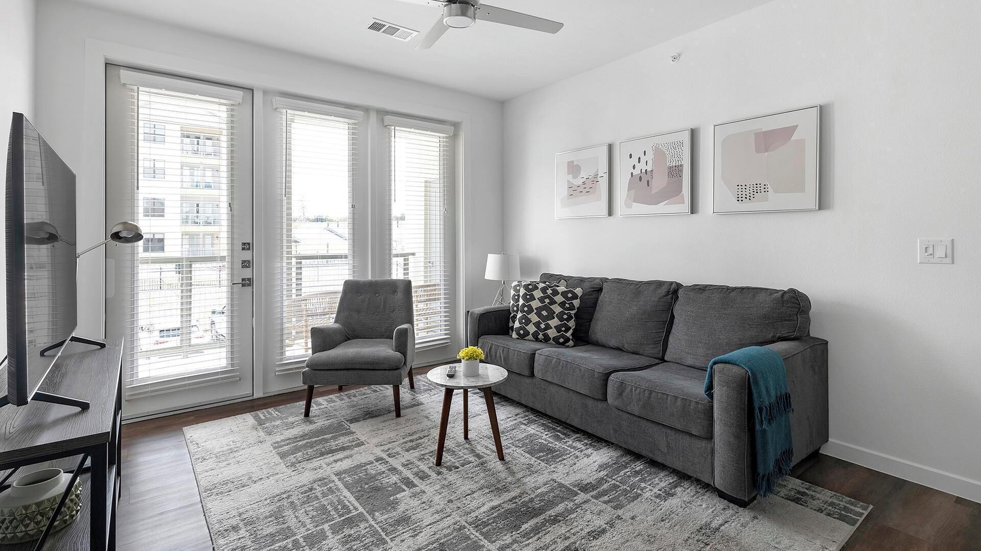 A living room with a grey sofa, a grey armchair, a round coffee table, and a TV on a stand. There are three framed artworks on the wall and a ceiling fan.