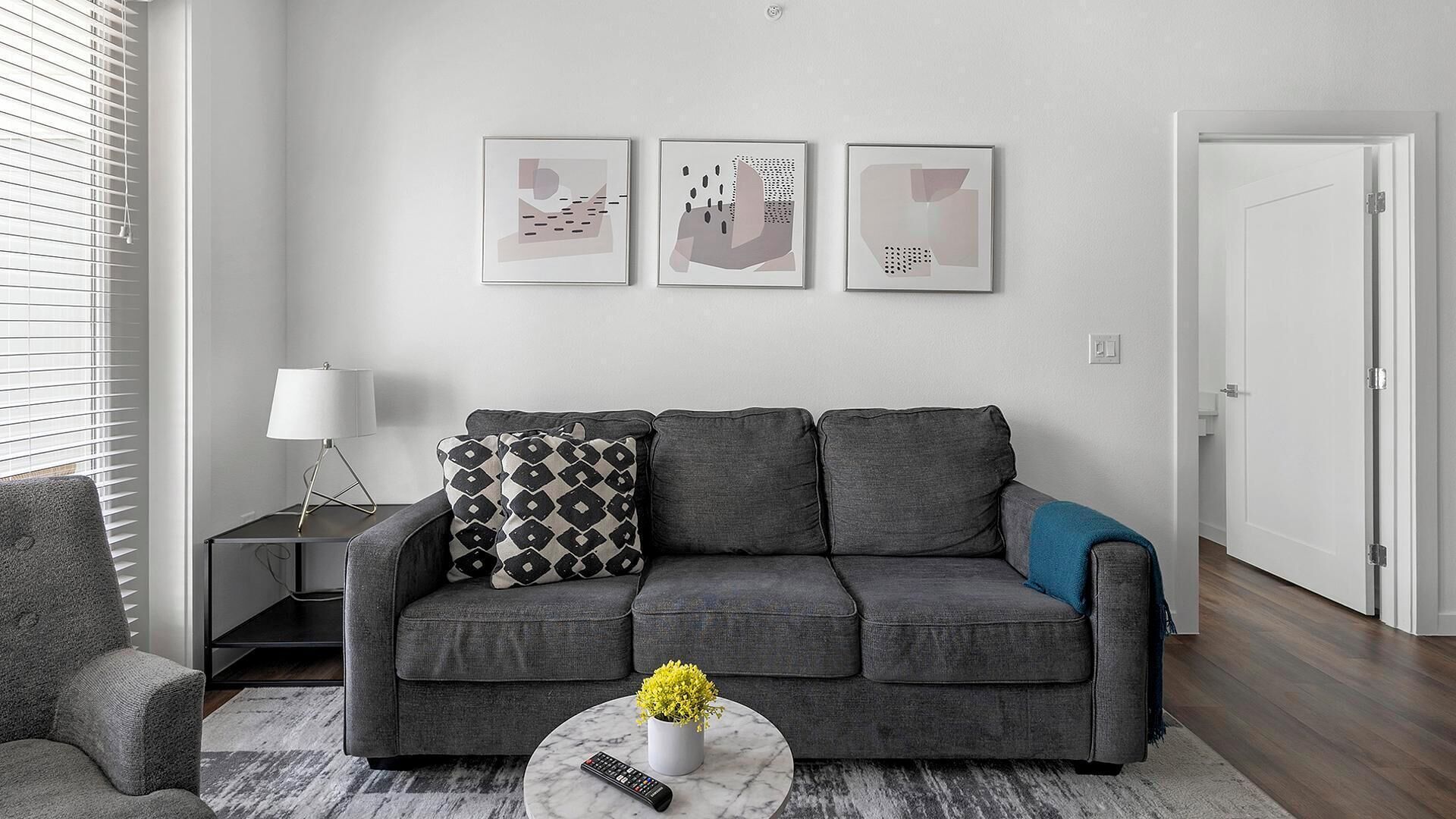 Ein modernes Wohnzimmer mit einem grauen Sofa, gemusterten Kissen, einer Fernbedienung und einer kleinen Pflanze auf einem Marmortisch. Darüber befinden sich drei abstrakte Wandbilder.