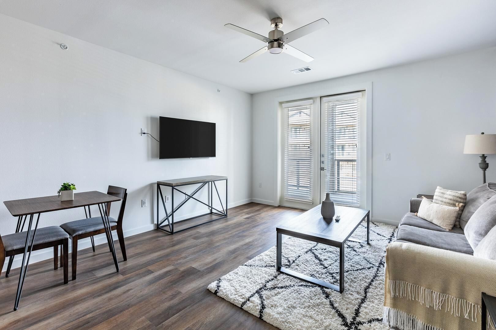 Un salon moderne comprenant un coin repas, un téléviseur à écran plat fixé au mur, un ventilateur de plafond et une grande fenêtre équipée de stores.