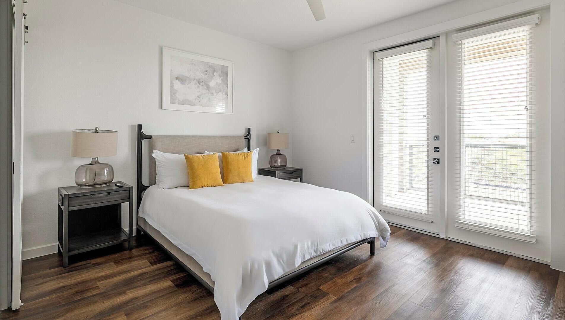 A bedroom with a bed, two bedside tables, a nightstand, a window with blinds, and a door.