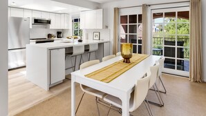 A modern kitchen with white cabinetry, a central island, and a dining area with a table and chairs. - Keith Haring Inspired 2BD Deauville (Palm Springs)