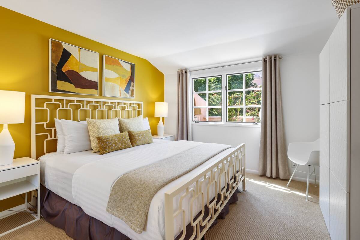 Une chambre moderne avec un grand lit, deux tables de chevet avec des lampes, une fenêtre avec des rideaux et des œuvres d’art abstraites accrochées au mur.