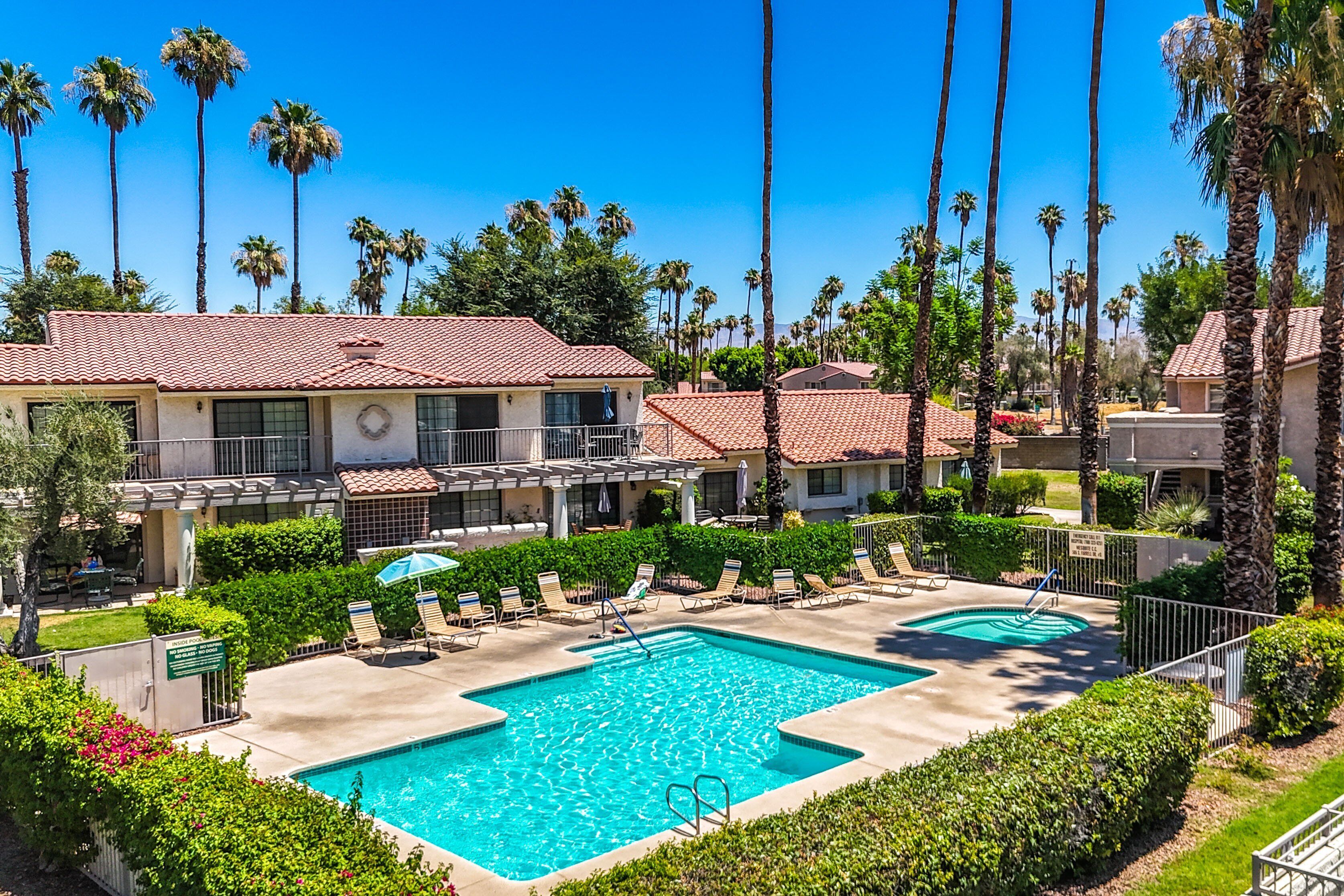 A pool area with lounge chairs and a swimming pool surrounded by palm trees and buildings with red-tiled roofs.