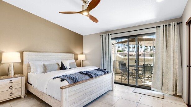 A bedroom with a bed, nightstands, a ceiling fan, and a view of the outdoors through a sliding glass door. - Quaint & Serene in Mesquite CC (Palm Springs)