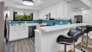 A modern kitchen with white cabinetry, a central island with bar stools, stainless steel appliances, and a blue backsplash. - Quaint & Serene in Mesquite CC (Palm Springs)