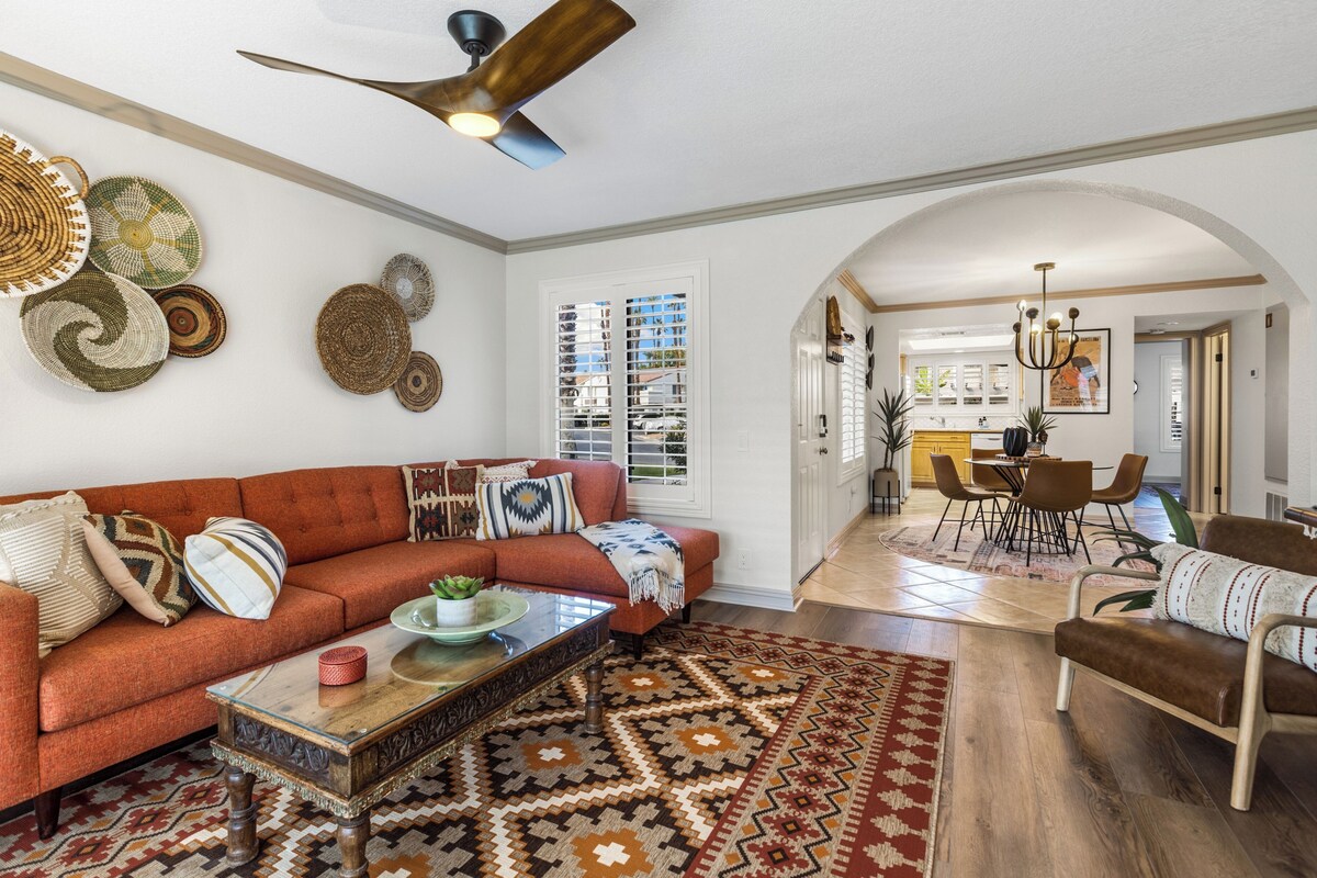 A spacious living room with a red sofa, a coffee table, and a patterned rug. There is a ceiling fan, a dining area with chairs, and a kitchen in the background.