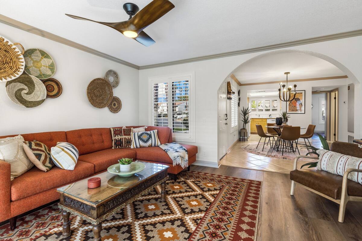A spacious living room with a red sofa, a coffee table, and a patterned rug. There is a ceiling fan, a dining area with chairs, and a kitchen in the background.