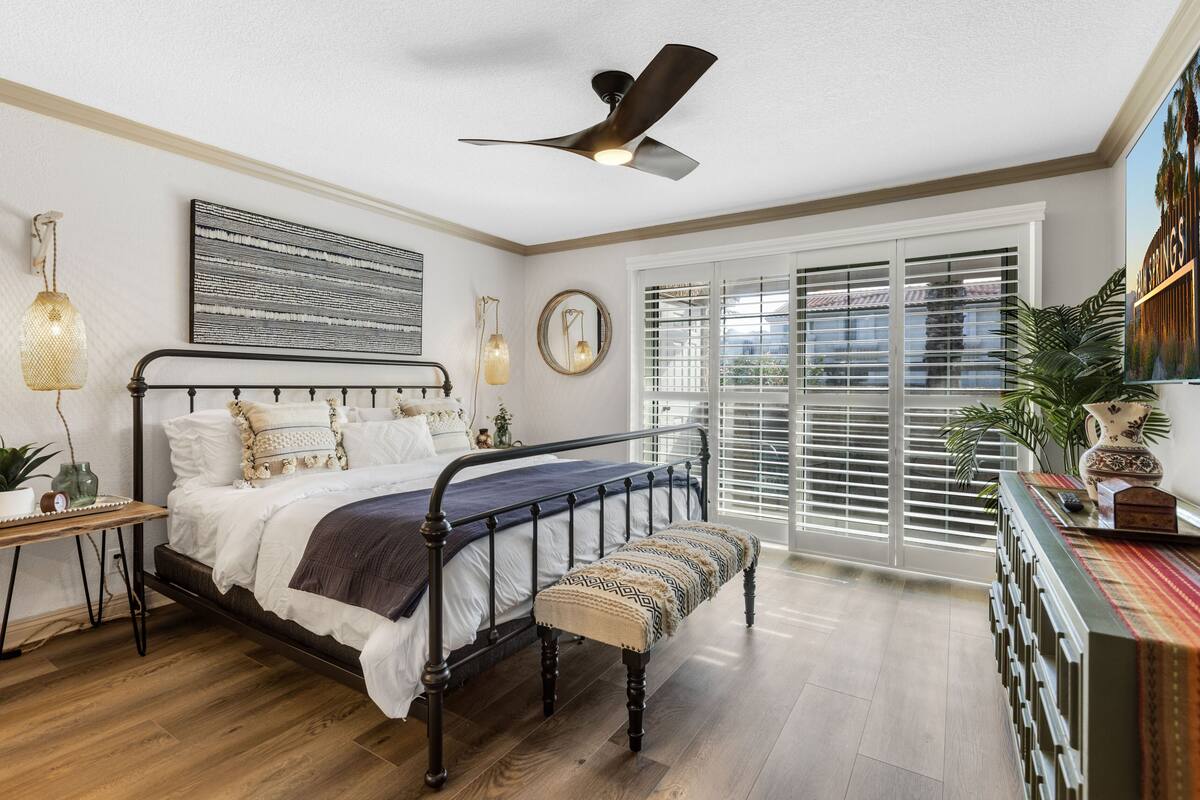 Une chambre à coucher comprenant un lit, une table de chevet, un banc, un ventilateur de plafond et une grande fenêtre avec des stores.